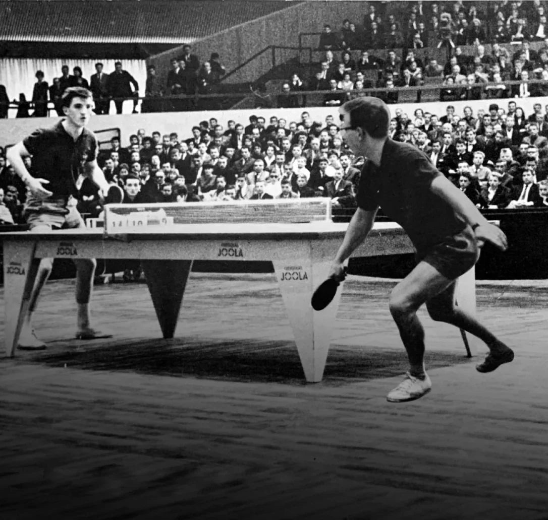 Two players compete in a table tennis match on a JOOLA table, with spectators watching in a vintage arena setting.